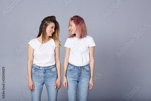 Horizontal portrait of two pretty women with happy expression, wears similar casual white t-shirt with denim, poses over gray background. Adorable lovely female student smile at each other. Copyspace