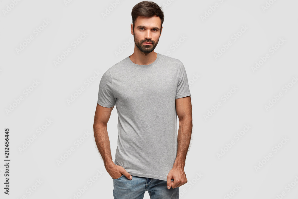 Fototapeta premium Stylish bearded handsome male with thick dark stubble, dressed in casual t shirt, keeps hand in pocket of jeans, looks self assured, fond of fashion, isolated over white background. Masculinity