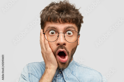 Omg, what I did! Surprised frightened young male with stubble touches cheek and opens mouth, being shocked from seeing how much he should pay in shop, stares through glasses, poses against white wall © Wayhome Studio