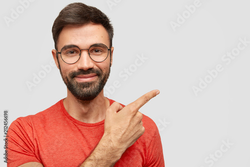 Photo of pleased unshaven European male with cheerful expression, has thick stubble, points aside, shows blank space for your advertising content, stands against white wall, dressed in red outfit