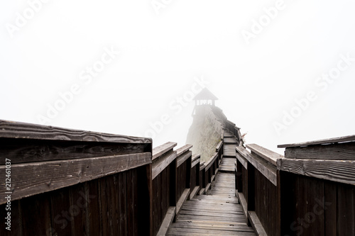 Wooden bridge leading to a tower on rocky ridge gradually disappearing in dense white fog. Boldogkovaralja, Hungary