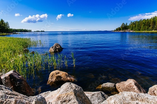 Konstfotografi Clear water blue lake with stones and green forest on a sunny summer day in Finland