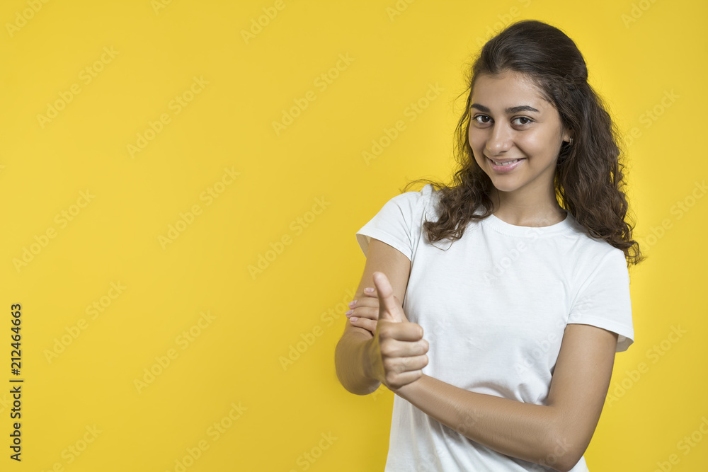 A joyful girl of mixed race holds her thumb up, against a yellow background. Copy space. A joyful girl of mixed race holds her thumb up, against a yellow background. Copy space.