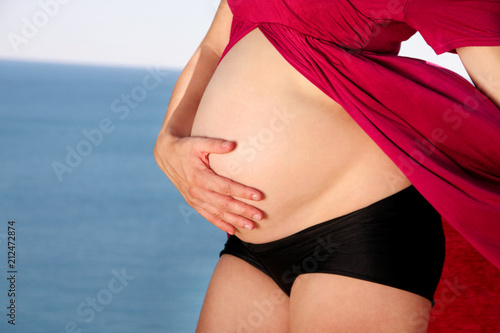 Beautiful pregnant woman and lady is hugging and holding her pregnant belly, standing in natural environment. Waiting for baby. Mom holds hands on belly on sea and sky nature background. Pregnancy.