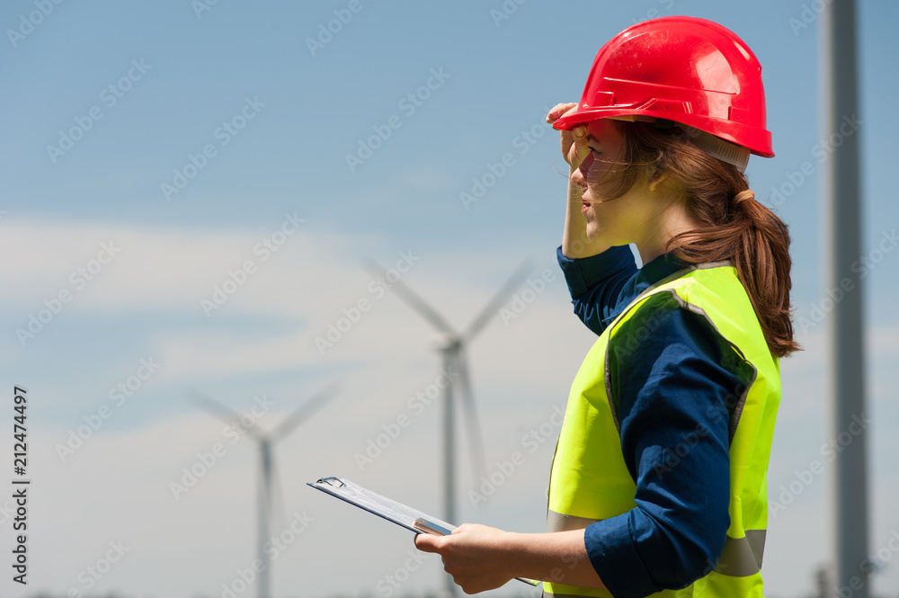 Concert of alternative energy and technology of the future. Side view cute young woman engineer in red helmet and green vest with work plan and smartphone in hands looking somewhere into the distance Concert of alternative energy and technology of the future. Side view cute young woman engineer in red helmet and green vest with work plan and smartphone in hands looking somewhere into the distance