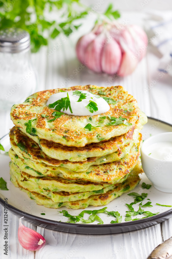 Zucchini pancakes with parsley and sour cream, summer food, tasty snack. High stack in a plate on white wood Zucchini pancakes with parsley and sour cream, summer food, tasty snack. High stack in a plate on white wood
