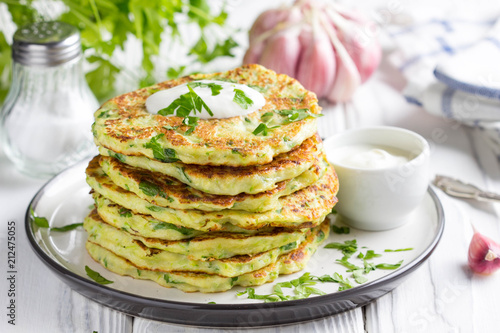 Zucchini pancakes with parsley and sour cream, summer food, tasty snack. High stack in a plate on white wood