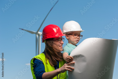 Concept of transfer of experience to the future generation. Woman engineer in red helmet shows her little son in glasses and white helmet work plan of a windmill