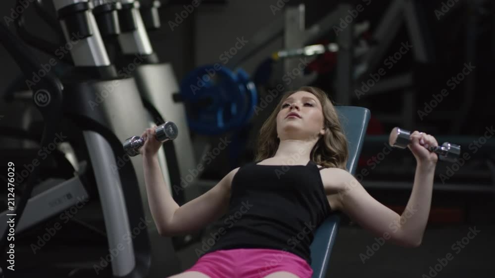 nice blonde curly girl with straight hair in a black sport wear with blue eyes does a dumbbell press on an incline bench and coach comes up to fix her close-up to face nice blonde curly girl with straight hair in a black sport wear with blue eyes does a dumbbell press on an incline bench and coach comes up to fix her close-up to face