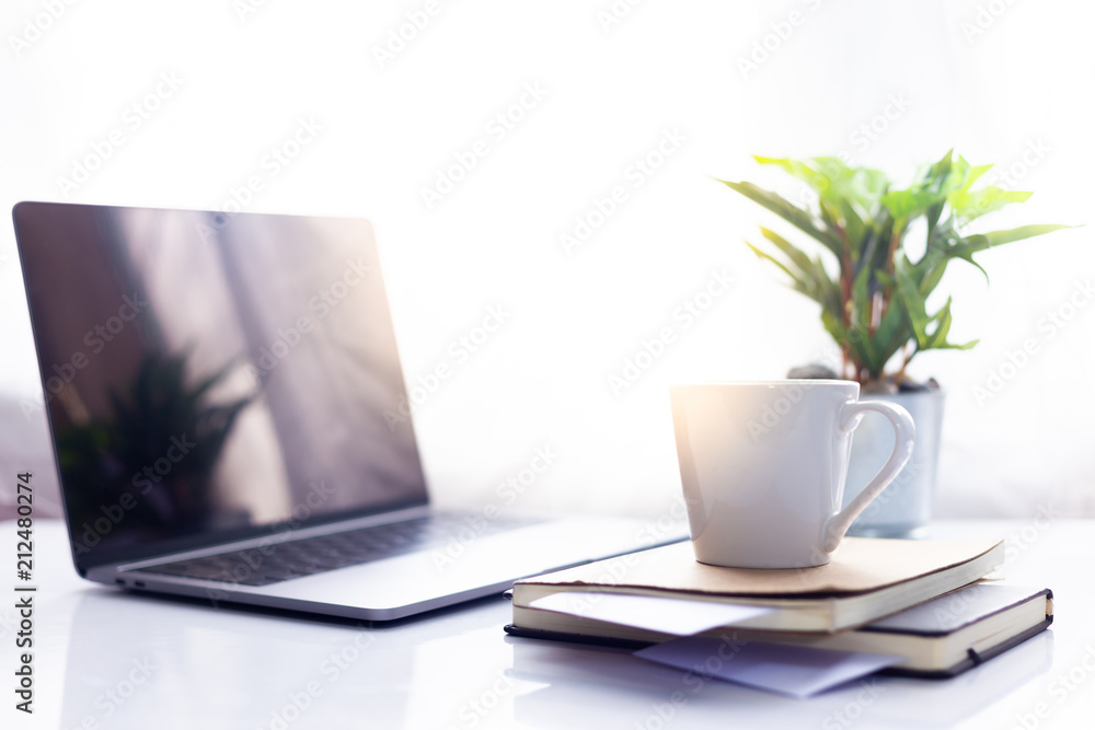 Coffee cup on notebook with laptop on white table in the morning sun light ,Working space Coffee cup on notebook with laptop on white table in the morning sun light ,Working space