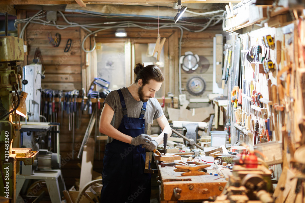 Young man in overalls standing by workbench and processing wooden workpiece Young man in overalls standing by workbench and processing wooden workpiece