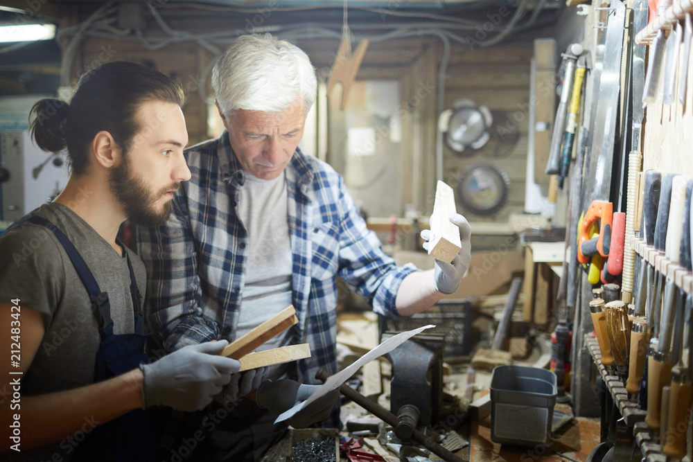Young trainee looking at wooden workpiece in hands of master while learning how to process wood Young trainee looking at wooden workpiece in hands of master while learning how to process wood