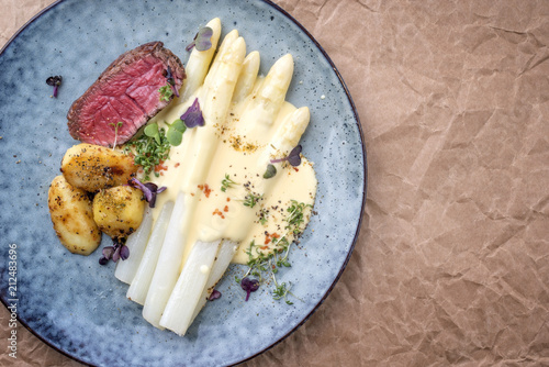 Modern barbecue dry aged sliced fillet steak with white asparagus in hollandaise sauce and roast potatoes as top view on a plate with copy space right