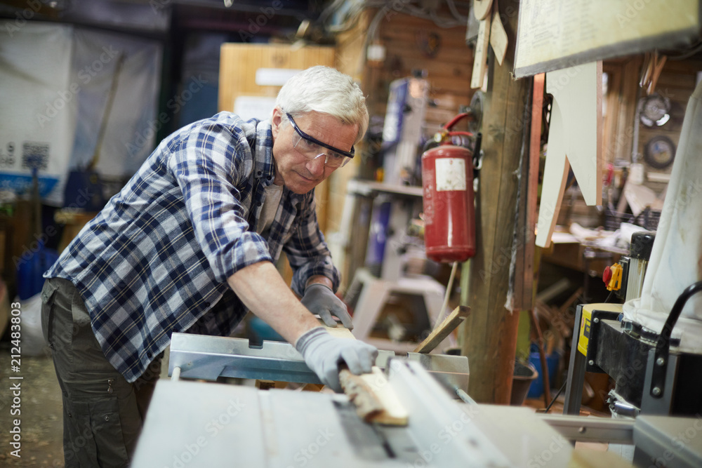 Aged master in protective eyeglasses processing wooden planks on machine in his workshop