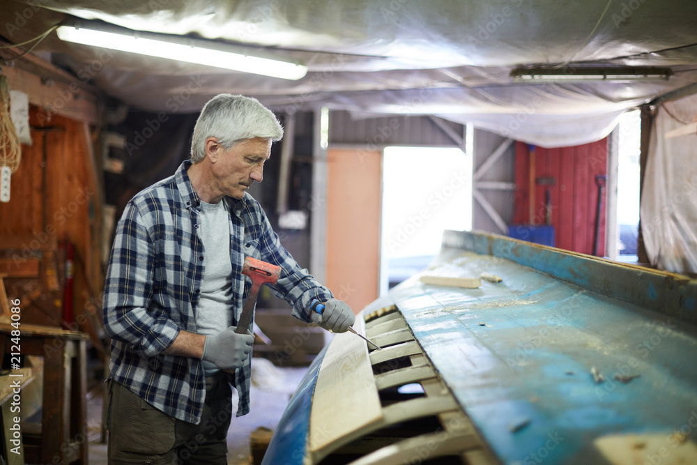 Aged master with chisel and hammer standing by vessel construction and processing its planks Aged master with chisel and hammer standing by vessel construction and processing its planks