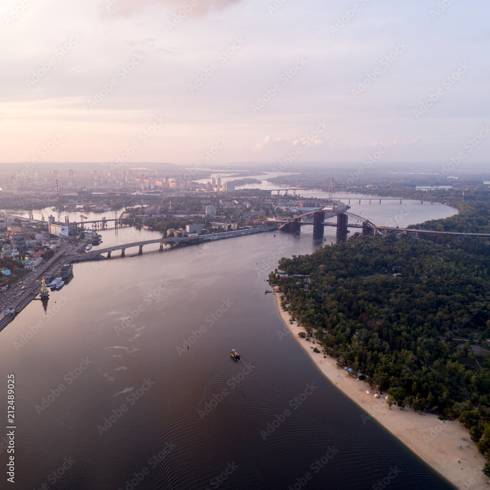 Fototapeta premium Panoramic view of a modern city with a river, unfinished bridge and park part of the city. Skyline bird eye aerial view with industrial zone and high-rise residential area under dramatic cloud sunset
