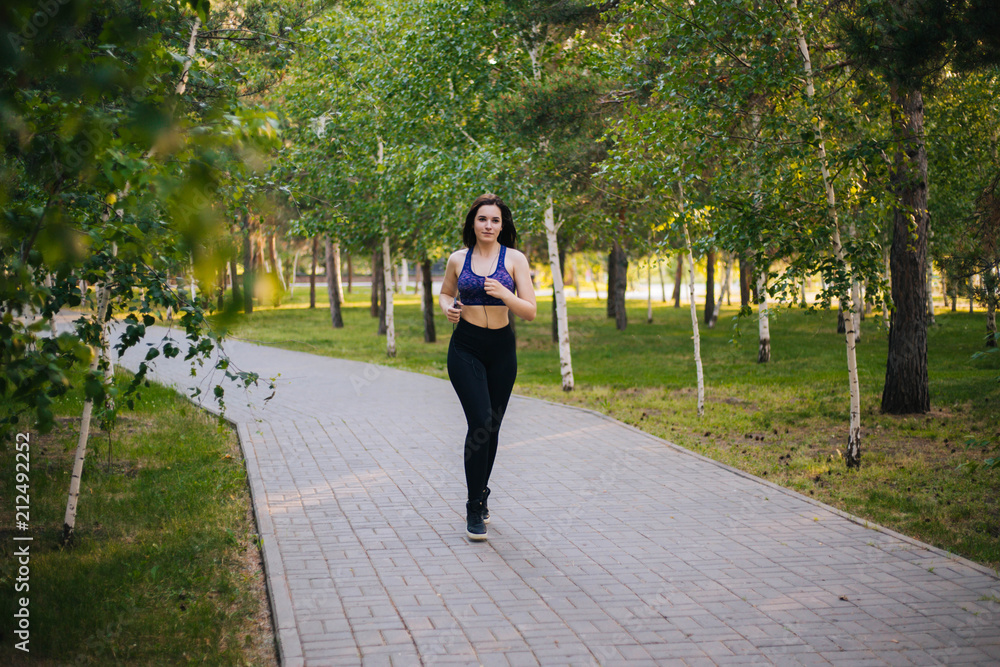 Young beautiful girl on a run. A woman is engaged in sports in the park. Healthy lifestyle. Running along the path with headphones, listening to music. Young beautiful girl on a run. A woman is engaged in sports in the park. Healthy lifestyle. Running along the path with headphones, listening to music.