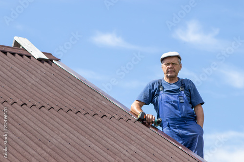 Old man working with electric screwdriver on a roof of a house with no safety harness, wearing work clothes, blue overall, dangerous job concept