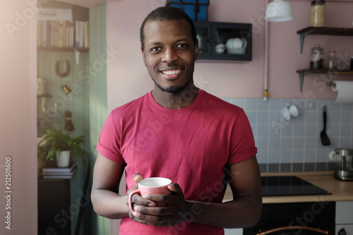 Indoor closeup photo of young African man standing in colored T-shirt in his kitchen smiling friendly, looking at camera, holding coffee cup in hands during his breakfast, enjoying free morning photo