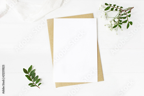 Feminine wedding stationery, desktop mock-up scene. Blank greeting card, craft envelope, baby's breath flowers, silk ribbon and lentisk branches. Old white wooden table background. Flat lay, top view.