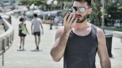 Young handsome man at the seaside using smartphone to record and send voice message, while looking at the beach and the sea