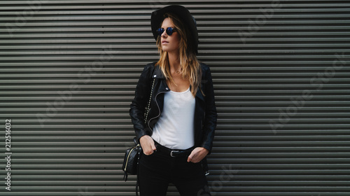 Stylish hipster girl with long blonde hair in sunglasses, wearing black jeans, leather jacket, sunglasses and hat looking aside while standing beside a gray wall with empty space for your content