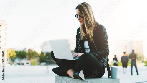 Young fashion blogger wearing stylish clothes writing new post in her blog by a laptop connected to wifi while sitting in a park with take away coffee in a paper cup on a warm sunny day.