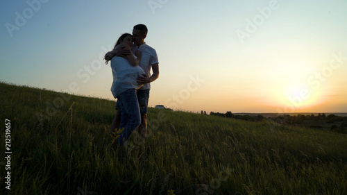 Romantic young happy pregnant couple hugging in nature at sunset. Man hugging woman from back in the evening in fields. Family, marriage, love, devotion, faith concept