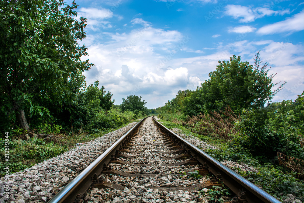 Railroad against beautiful blue cloudy sky in nature. Railway tracks through the woods at day. Transport and shipping landscape concept