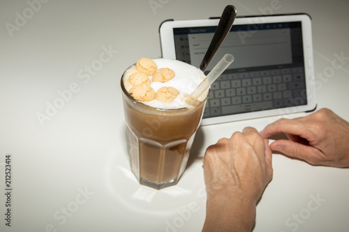 Person schreibt auf Tablet. Neben dem Tablet steht ein Glas mit Eiskaffee. Oben ist aufgeschäumte Milch und einige Amaretti. Außerdem ist ein durchsichtiger Strohhlam und ein langer Löffel im Glas. photo