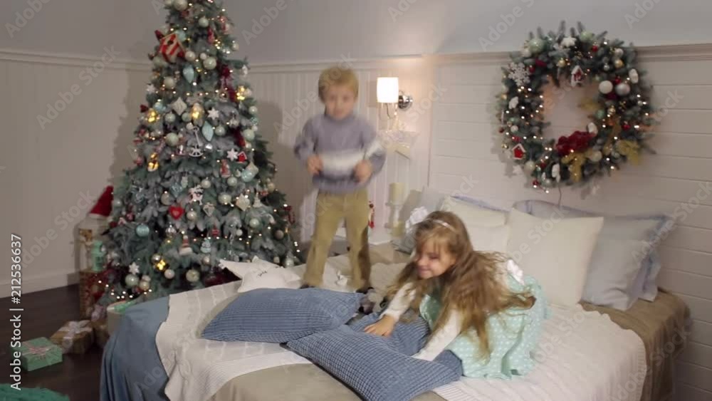 Two young happy cheerful children jumping on the bed at Christmas with a Christmas tree. Two young happy cheerful children jumping on the bed at Christmas with a Christmas tree.