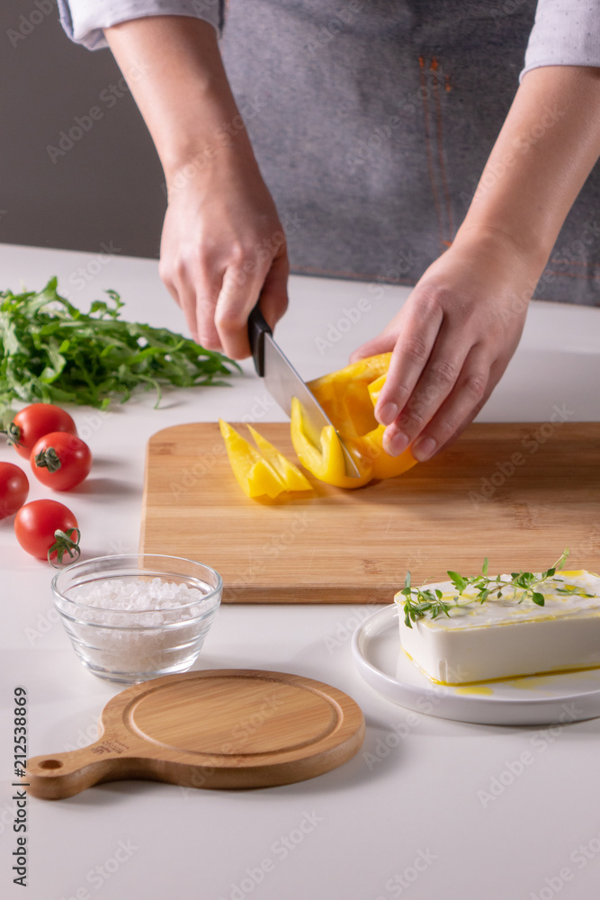 The girl's hand cuts pieces of pepper on a wooden board, cheese, and tomatoes on a white kitchen table. Preparation of salad The girl's hand cuts pieces of pepper on a wooden board, cheese, and tomatoes on a white kitchen table. Preparation of salad