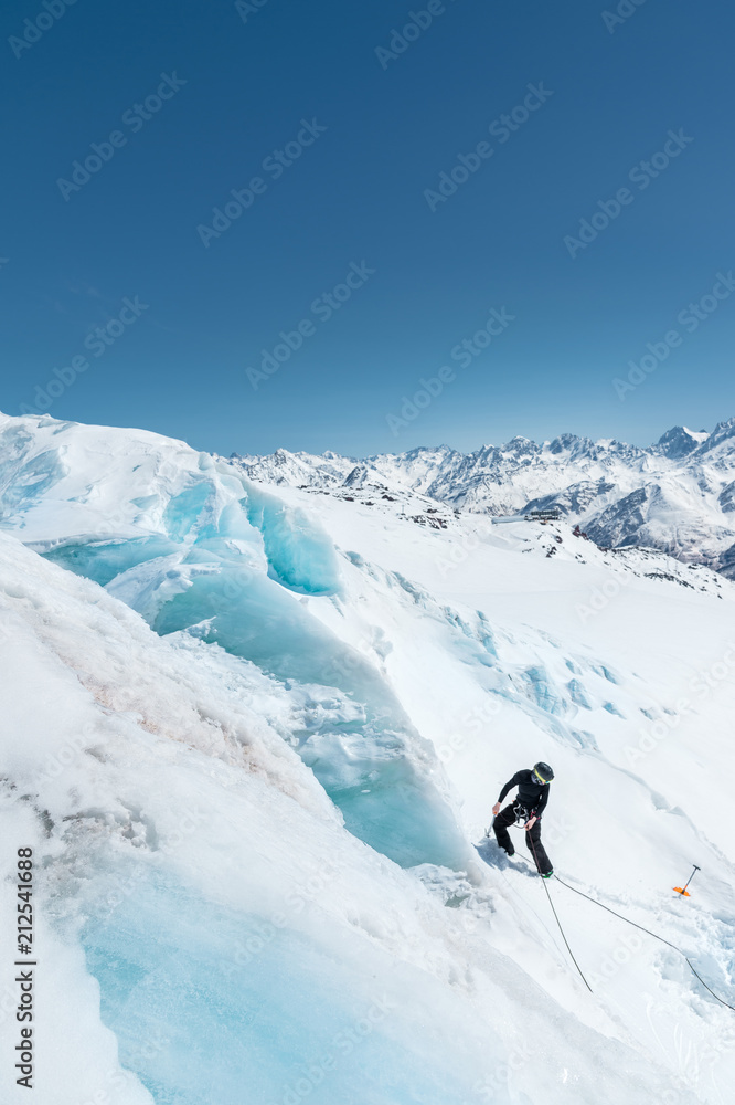 A professional mountaineer in a helmet and ski mask on insurance makes a nick-hole in the glacier against the backdrop of the Caucasian snow-capped mountains