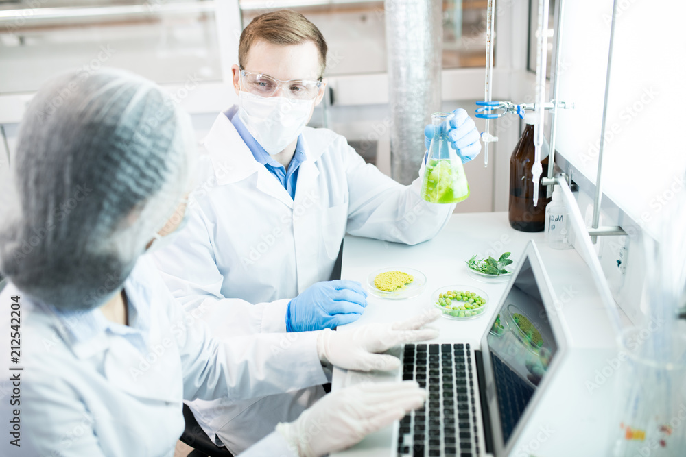 Portrait of two modern scientists working on food research studying liquids in beaker while sitting at table in laboratory Portrait of two modern scientists working on food research studying liquids in beaker while sitting at table in laboratory