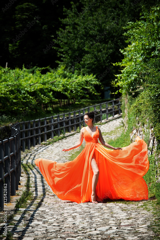 Beautiful girl in amazing fluttering orange dress standing on the cobblestone curved road. Beautiful girl in amazing fluttering orange dress standing on the cobblestone curved road.