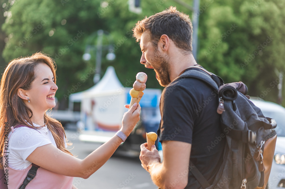 Naklejka premium happy young woman feeding boyfriend with closed eyes by ice cream at urban street