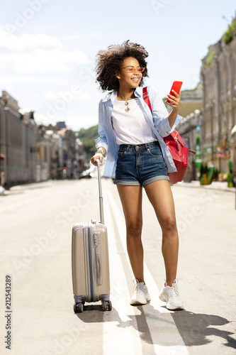 Just arrived. Pleasant female tourist using her phone and searching for a hotel location while carrying her suitcase