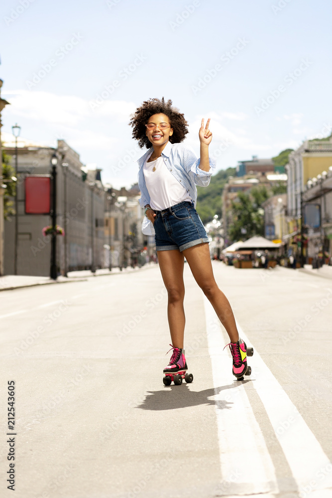 Cute teen. Pleasant curly girl posing for the camera and showing V sign while roller-skating in the streets Cute teen. Pleasant curly girl posing for the camera and showing V sign while roller-skating in the streets