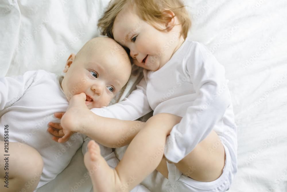 Two charming little babies lie on the white sheet and smile Two charming little babies lie on the white sheet and smile