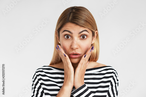 Horizontal shot of attractive bug eyed young blonde Caucasian woman wearing stylish striped t-shirt keeping hands on cheeks and raising eyebrows, shocked with big sale prices or unbelievable gossip