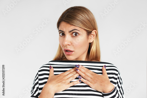 Isolated portrait of astonished young woman with blonde hair reacting emotionally, startled with unexpected sudden noise, holding hands on chest, her look expressing shock, fright or terror