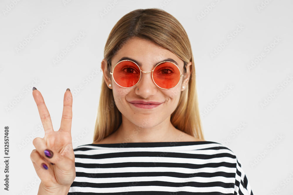 Beauty, style, glamour and fashion concept. Charming stylish young blonde female wearing striped top and trendy pink sunglasses in round shape smiling happily, showing peace sign at camera Beauty, style, glamour and fashion concept. Charming stylish young blonde female wearing striped top and trendy pink sunglasses in round shape smiling happily, showing peace sign at camera