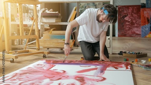 Enthusiastic bearded painter making a picture in art studio, dealing with huge work of art on wooden floor, wearing dark jeans and white t-shirt, indoor shot on beautiful sunny day