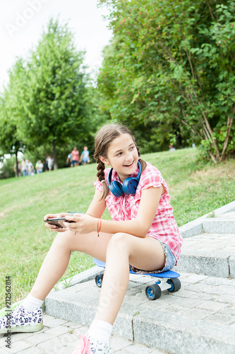 Girl having fun with her phone in the park while sitting down on her skateboard 
