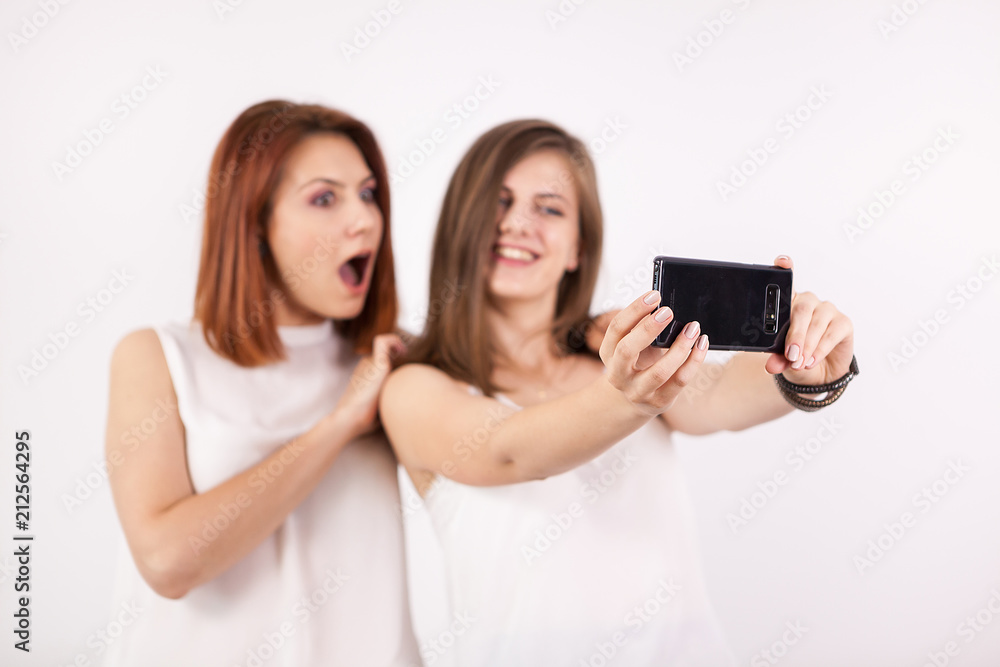 Two happy girls taking a selfie in studio on a white wall Two happy girls taking a selfie in studio on a white wall