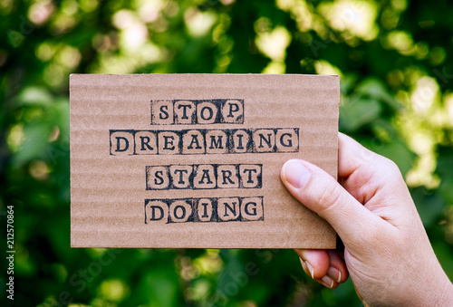 Woman hand holding cardboard card with words Stop Dreaming Start Doing against green nature background. © rosinka79