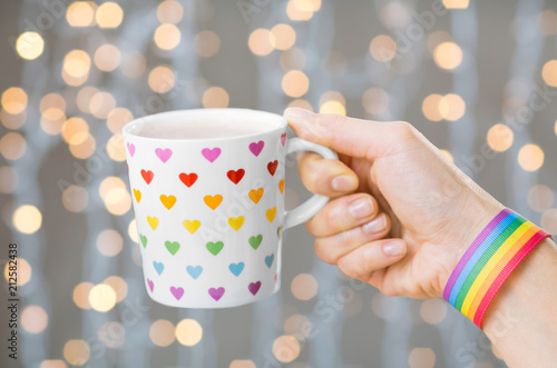 homosexual and lgbt concept - close up of female hand with cacao in cup with heart pattern and gay pride awareness wristband over festive lights background