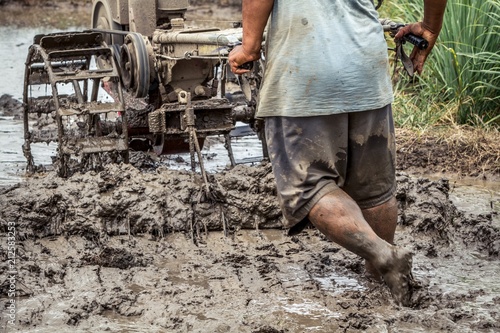 Strong asian farmer driving tiller tractor in muddy field, detail of male farmer walking barefoot in deep mud, hard work in field