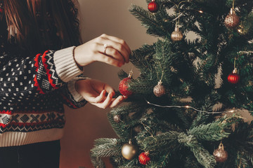 woman in stylish sweater decorating christmas tree in room. merry christmas and happy new year concept. space for text. woman putting on red ornaments. family cozy moments