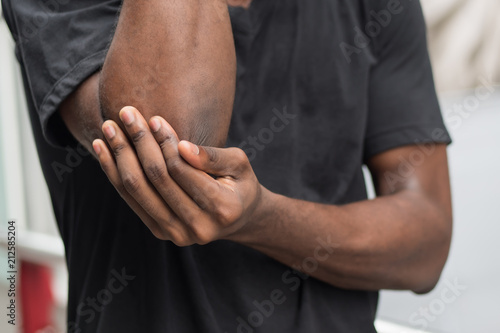 african man suffering from elbow pain; sick black man with elbow joint pain, osteoporosis, bone arthritis, gout, symptoms; body sickness, health care or pain concept; adult african man or model